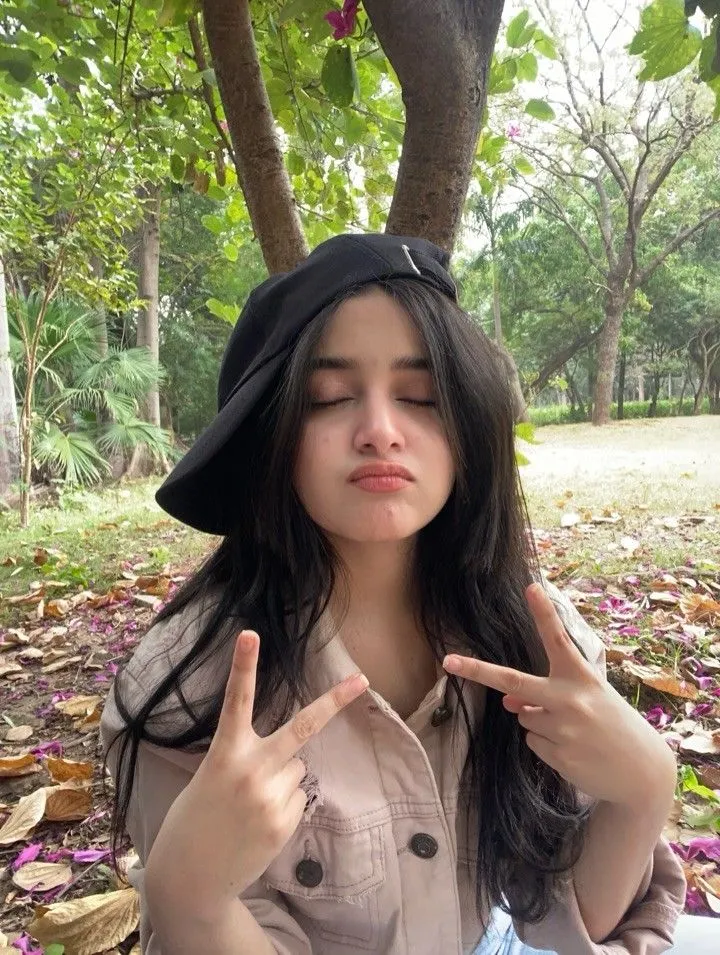 a woman sitting under a tree making the peace sign with her hands while wearing a hat