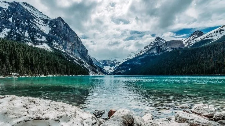 a lake surrounded by snow covered mountains under a cloudy sky with clouds in the distance