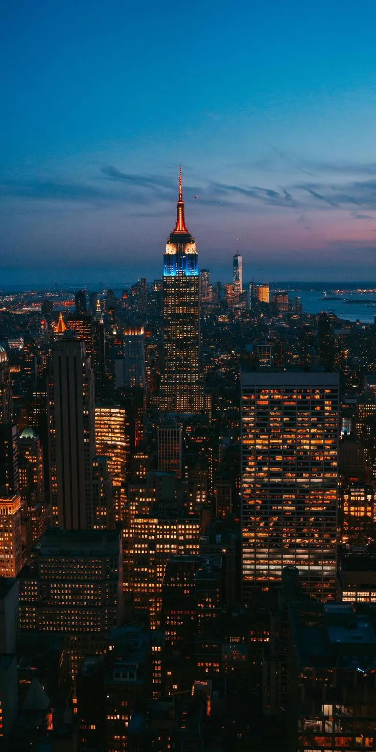 the empire building is lit up at night in new york city, with skyscrapers visible