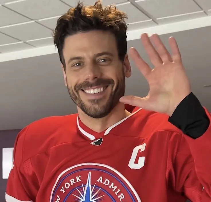 a man in a red hockey jersey waving at the camera with his hand up and smiling