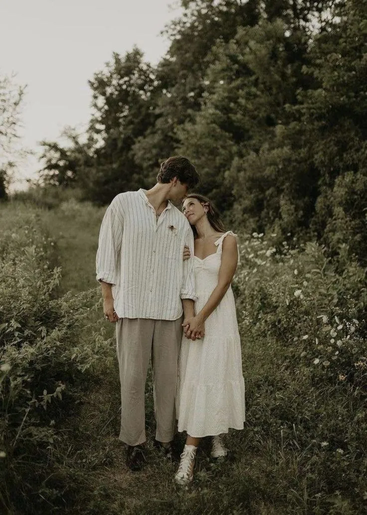 a man and woman standing next to each other in the grass with trees behind them